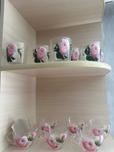 A set of clear glass drinking vessels with hand-painted pink roses and gold accents are displayed on two wooden shelves. The upper shelf holds four glasses, each decorated with a rose motif and a textured, diamond-like pattern on the glass. The lower shelf displays five teacups, also featuring hand-painted roses. One teacup on the lower shelf has a piece of paper tucked inside it. The shelves are part of a light-colored wooden cabinet, with the back of the cabinet and a gray wall visible behind the shelves. The location is Yazykova Pyatina, Russia.
