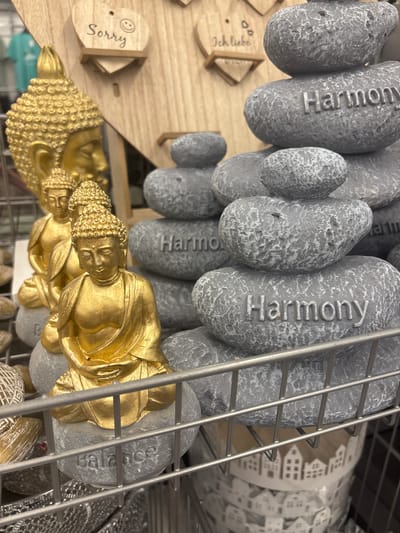 This media file displays a close-up, angled view of various decorative items, likely intended for home decor or spiritual practice. The primary subjects are several large, gray, stone-like objects with the word "Harmony" inscribed on them, arranged vertically. In the foreground, a gold-colored Buddha statue is prominently featured, also angled and partially obscured by the gray stones and a metal grate. Another, smaller gold Buddha figure is visible behind the first.
To the left, a small wooden heart-shaped object with a smiley face and the German text "Ich liebe" (I love) is visible. Further down, another wooden heart-shaped object reads "Sorry" in cursive. The background shows a roll of paper or fabric with a repetitive pattern of small white houses.
The setting appears to be a retail environment, possibly a store display, given the metal grate and the arrangement of merchandise. The lighting is artificial, and there are no indications of time of day or weather. The overall impression is one of calm and mindfulness, suggested by the "Harmony" inscriptions and the Buddha imagery. The text "Ich liebe" and "Sorry" add a personal, emotional touch to the display. The location context indicates this is Erlenbach am Main, Germany.
