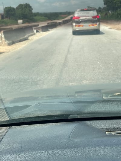 The image shows a view from inside a vehicle, looking out through the windshield at a road under construction. A light-colored SUV is in front, driving away from the camera. The road appears to be paved and is lined with concrete barriers. In the background, there are trees and a cloudy sky. The scene suggests a rural or developing area, possibly on the outskirts of Aciol, Nigeria. The construction on the road indicates ongoing infrastructure development.