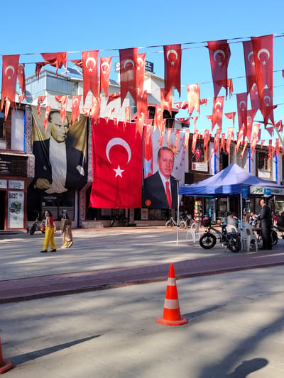 This image captures a lively street scene in Iznik, Turkey, under a clear blue sky. Numerous red Turkish flags, adorned with a white crescent moon and star, are strung across the street, creating a festive atmosphere.

Three large banners dominate the upper part of the street. On the left, a banner displays a portrait of Mustafa Kemal Atatürk, the founder of the Republic of Turkey, dressed in a suit. Next to it, a large Turkish national flag hangs vertically. On the right, another banner features a portrait of President Recep Tayyip Erdoğan, also in a suit and red tie, with a slight smile.

At street level, two individuals, possibly children, are walking past the Atatürk banner. One wears bright yellow traditional attire, while the other is dressed in a striped garment. Further down the street, a blue and purple canopy tent is set up, under which several people are gathered at tables, appearing to socialize or dine. A man in a dark suit stands near the tent. Motorcycles and scooters are parked in the vicinity, and white plastic chairs are visible.

In the foreground, an orange and white traffic cone stands on the paved ground, casting a long shadow, indicating a sunny day. The surrounding buildings house various establishments. Visible text on signs includes "IZNIK," "ZABITA NOKTASI" (Municipal Police Point), "EL MEDYA," and "TURKIYE," providing specific location and contextual information. The scene depicts a bustling public space, possibly decorated for a national holiday or event.