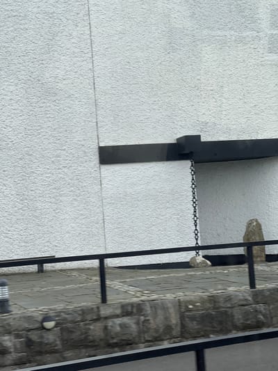 The image shows the exterior of a building in Creeslough, Ireland. A textured white wall dominates the background, with a dark horizontal beam extending from the upper right of the frame. Hanging from this beam by a chain is a rock. To the right of the hanging rock, a larger, rough-hewn stone stands upright. In the foreground, a black metal railing runs horizontally across the scene, separating a paved walkway from a stone wall below. The walkway is made of grey flagstones and cobblestones. A cylindrical light fixture with silver and grey elements is visible on the left side of the walkway.