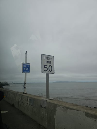 A speed limit sign reading "SPEED LIMIT 50" is mounted on a metal pole. To the left of the speed limit sign, a blue and white sign reading "CALL BOX" with "AL 80 15" underneath is also mounted on a pole. The signs are located on the side of a road next to a body of water. A concrete barrier runs along the edge of the road. The sky is overcast and the water appears choppy. The scene is likely located in Oakland, California, United States of America.