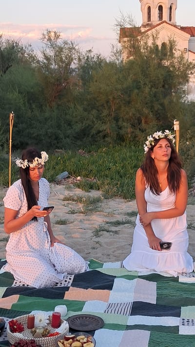 In Agia Triada, Greece, two women are seated on a picnic blanket on a sandy beach. The woman on the left, wearing a white polka-dot dress and a floral crown, is looking at her phone. The woman on the right, in a white dress and a floral crown, is looking to the side. Behind them, dense green foliage grows, and a white building with a domed roof is visible in the distance. A bamboo torch is placed to the left of the woman on the left, and a picnic basket with fruit, candles, and flowers is in the foreground.