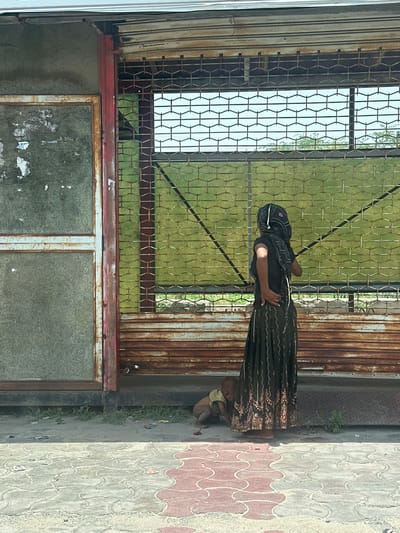 The image captures a scene outside what appears to be a shop or establishment in Ajmer, India. A woman, dressed in a dark, patterned skirt and a headscarf, stands with her back to the viewer, facing a metal gate. Her left hand is on her hip. To her left, a young child is lying on the ground, partially obscured by the woman's skirt. The child is wearing a light-colored garment. The environment shows a concrete walkway with a red and gray patterned surface in the foreground. Behind the woman and child is a rusty metal gate with a green mesh screen behind it. To the left of the gate, there is a section of a building with a faded, grimy window and a red metal frame. Debris, including small colorful objects, is scattered on the ground near the building and gate. The overall impression is one of a somewhat neglected urban environment.