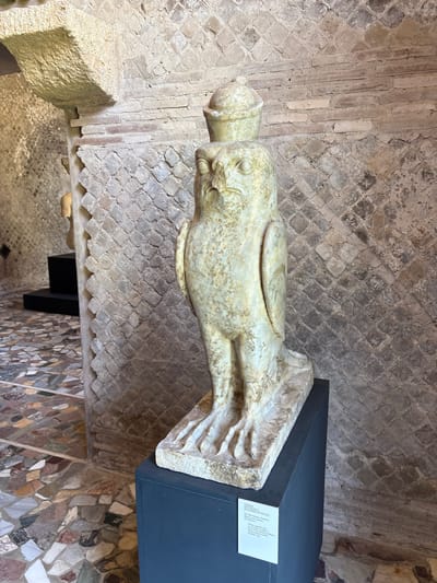 The image shows a marble sculpture of a falcon, identified by a nearby sign as a "HORCIO IN FORMA DI FALCO," found in the Palestra of Villa Adriana in Tivoli, Italy. The sculpture is made of Proconnesia marble and depicts the Egyptian god Horus in the form of a falcon. The sculpture rests on a modern dark gray display platform. The background shows remains of a wall built with a herringbone brick pattern. The floor is covered with a fragmented mosaic floor.