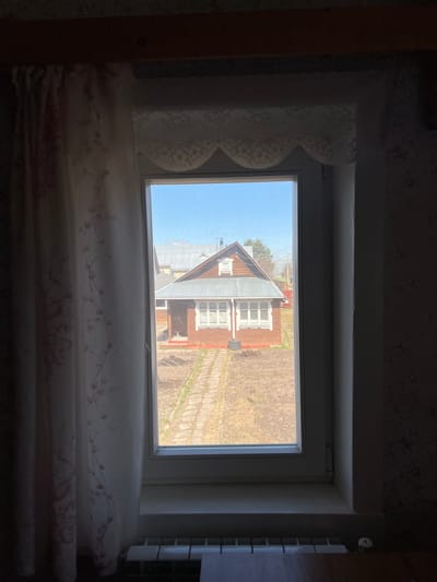 The image depicts a view from a window of a house in Чернышово, Russia. Outside the window, another house with a grey roof and brown siding is visible, as well as bare patches of ground. A faint walkway leads to the house. The sky is light blue. Inside, the window is draped with patterned curtains, and a radiator can be seen on the right. The overall scene appears to be taken during the day, likely early spring or late fall.