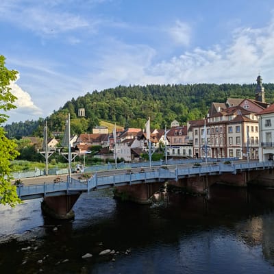 The image shows the Murg River flowing through the city of Gernsbach, Germany. A bridge crosses the river, with buildings lining the far bank. The bridge is made of metal and has a blue railing. There are two people standing on the bridge, looking out at the river. The buildings on the far bank are a mix of old and new, with some traditional half-timbered houses and some more modern apartment buildings. A dense, green forest covers the hillside behind the buildings, and the sky is a clear blue with a few wispy clouds.