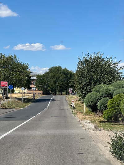 The image depicts a rural road curving to the right, bordered by lush green trees and vegetation on the left and a sidewalk and more greenery on the right. The sky is a clear, bright blue with a few scattered white clouds, suggesting a sunny day, likely mid-morning or afternoon due to the strong sunlight and shadows.
Along the roadside, several signs are visible. One sign, a red and white circular speed limit sign, indicates "70". Another circular blue sign displays an icon of a pedestrian and text. A row of directional signs, with red and white lettering, point towards various locations including "Kostice," "U Votrolamu," "Polni," and "Hřbitov" (which translates to Cemetery). In the background, a building with a yellow facade and several windows is visible, partially obscured by trees. The overall scene conveys a tranquil, everyday setting in a Czech village. No specific activity or event is clearly depicted, beyond the general scene of a road in a populated area.