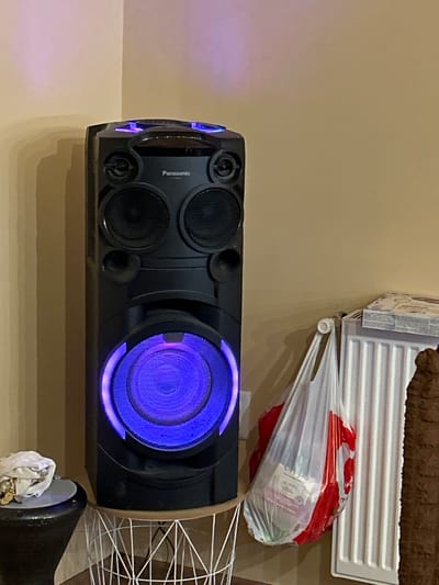A tall, black Panasonic speaker with blue lighting is positioned in the corner of a room, adjacent to a white ribbed radiator. A white plastic shopping bag containing red and green items hangs from the radiator. To the left of the speaker, a black, cone-shaped object topped with a grey circular surface holds a bundle of white fabric and several gold-colored objects. The wall behind the speaker and radiator is a light tan color. The floor is not visible.
