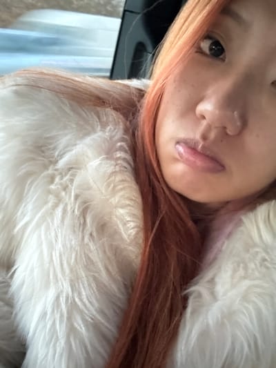 A close-up shot features a woman with vibrant red-orange hair, freckles, and glossy lips. She's nestled against a fluffy, white faux fur coat or blanket, creating a soft and warm atmosphere. A sliver of a dark object appears on the left edge. The image is angled, offering an intimate perspective of the subject's face and texture.