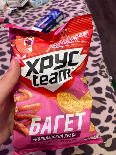 A hand is holding a brightly colored snack bag featuring "ХРУСТ team" and "БАГЕТ" in large white lettering against a pink and red background. Images of crab legs and toasted bread slices are also visible. The background appears to be a patterned fabric, possibly a blanket or bedding, with a blurred assortment of items behind it, including what appear to be cigarette packs and a blue tube. The image does not contain any identifiable landmarks or contextual clues to suggest a specific location within the city of Vaganovo, Russia.