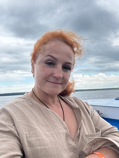 A woman with red curly hair and fair skin, wearing a tan button-up shirt, takes a selfie on a boat. She is smiling slightly and looking at the camera. Her hair is windswept. She is wearing a black cord necklace. In the background, a body of water is visible with a treeline on the horizon under a cloudy sky. The foreground shows the railing of the boat and a blue surface, possibly water. The location is Ikskoe Ust'e, Russia.