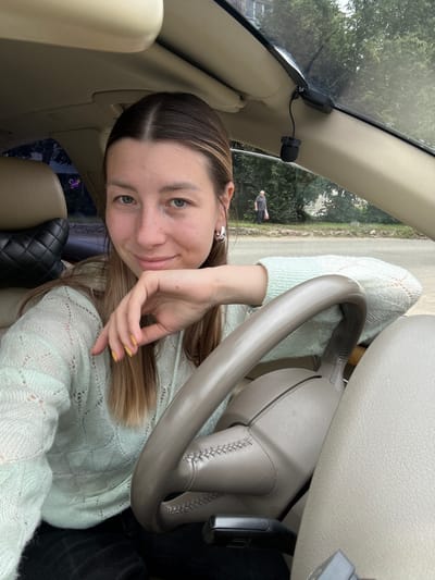 A young woman with light brown hair is sitting in the driver's seat of a car. She is wearing a light green sweater. Her left arm is resting on the steering wheel, and she is looking at the camera. The interior of the car is visible, including the dashboard, steering wheel, and seats. Outside the car, there are trees and a building. A man is walking in the background. The image appears to have been taken in Yartsevo, Russia.