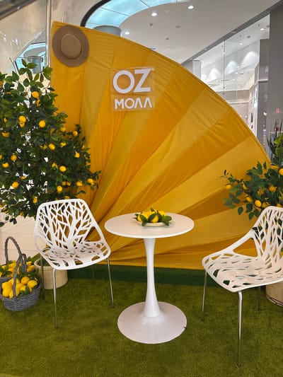 The image shows an outdoor seating area with a yellow fan-shaped backdrop, possibly at a mall in Tlustenkhabl, Russia. Two white modern chairs are placed on green artificial grass, facing a small white table. The table has a bowl of lemons on it. To the left of the chairs, a lemon tree with artificial lemons is visible, and next to it is a basket filled with lemons. A brown hat is hanging on the yellow backdrop. The background shows a bright, modern interior with glass walls and ceiling lights, consistent with a shopping mall environment. There are no people present in the image.