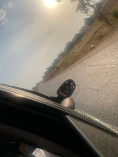 The image presents a tilted, outdoor scene captured from the perspective of someone inside a moving vehicle, looking out the side window. If oriented correctly (rotated approximately 90 degrees counter-clockwise), the road would run horizontally across the lower part of the frame, and the sky would be at the top.

In the foreground, occupying the right side of the image, is the dark frame of a car's door or window, along with the car's side-view mirror. The mirror casing is dark, and its glass reflects a blurred image, likely of the car's interior or the road behind. An amber turn signal indicator light is visible on the side of the mirror assembly.

Outside the car, a light-colored asphalt road stretches diagonally across the middle of the frame (horizontally if rotated). The road surface shows visible cracks and some light wear. To the left of the road is a dry, earthy shoulder or verge, with patches of light brown grass and some greener vegetation, suggesting a somewhat arid or rural environment. Further in the distance, a line of trees or dense bushes forms a natural horizon line, extending across the mid-ground.

The sky dominates the upper and left portions of the image (or upper-left if rotated). It is very bright and appears hazy, with a strong, low-lying sun visible in the upper-left corner (if rotated). This sun creates a significant glare, washing out part of the sky and bathing the scene in a warm, yellowish-white light. A few faint, scattered clouds are discernible in the less glary areas of the sky.

The strong, low sun indicates either early morning or late afternoon/evening. The overall atmosphere is bright and hazy, suggesting clear weather with possibly some atmospheric dust or moisture contributing to the haze. The scene depicts travel, likely along a rural or semi-rural road. No text is visible in the image.