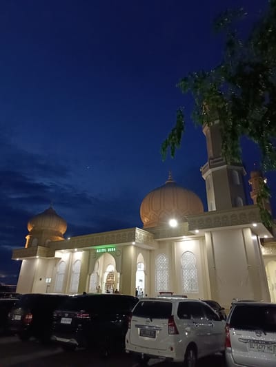The image captures an exterior view of the Baitul Musa Mosque in Mee Tanjong Usi, Indonesia, at twilight. The mosque is a prominent structure, featuring two large domes, one more prominent than the other, and a tall, slender minaret. The building's facade is illuminated, highlighting decorative architectural elements and intricate window designs. The sky above is a deep blue, with wispy clouds and a faint, distant light, possibly a planet or star. In the foreground, several vehicles are parked, including SUVs and sedans, suggesting this is a public gathering place or a parking area adjacent to the mosque. The overall scene conveys a sense of calm and reverence, typical of a religious site during evening hours.