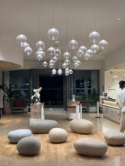 This image captures a modern interior space, identified as a lobby or reception area, located in Marcq-en-Barœul, France. The scene is illuminated by numerous clear, spherical glass pendant lights of varying sizes suspended at different heights from a white, perforated acoustic ceiling.
The floor consists of light-toned wooden planks arranged in an irregular pattern. Six large, oval-shaped poufs, upholstered in shades of grey, beige, and light blue-grey fabric, are positioned in the foreground and midground.
In the midground-left, the partial back of an individual in light clothing is visible. Further back, a dark grey armchair with a red cushion is situated next to a potted palm plant. Centrally, two slender, slatted wooden pedestals are present; one displays a white floral arrangement. Behind these elements, large glass doors, consistent with a revolving or sliding entrance, open to a dark exterior or another internal space, suggesting evening or night conditions.
To the right, a reception desk constructed from light wood and white textured panels features various items on its surface. Above the desk, a white wall displays a black arrow pointing left and the word "SANITAIRES" in black capital letters. A male individual, viewed from the back, wearing a light t-shirt, light pants, and a dark baseball cap, stands in front of the reception desk. Another potted palm is located behind the desk. Additional spherical pendant lights similar to the main ceiling cluster hang above this area.