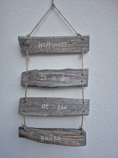 The image shows a rustic wooden sign hanging on a textured white wall. The sign is made of four pieces of weathered, grey wood, arranged vertically and strung together with twine. Each piece of wood has white lettering painted on it, forming the phrase "HAPPINESS IS a day at the beach". The wood has knots and visible grain, suggesting it may be driftwood. The wall appears to be an exterior wall, possibly of a building. The lighting suggests it is daytime, with diffuse light casting no strong shadows. There are no people or animals visible in the image. The background is entirely the textured white wall. The image does not contain any information or visual cues that would allow for its location to be identified as Hagen, Germany.