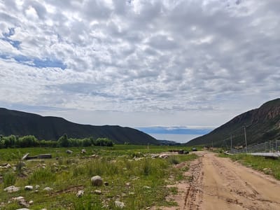 A wide-angle, eye-level shot captures a natural landscape dominated by rolling hills and a dramatic sky. A dirt road, with visible tire tracks, occupies the foreground on the right, leading towards the distant horizon. The road is flanked by sparse green grass and scattered rocks, with a denser patch of green vegetation and trees on the left. In the distance, a body of water, possibly a lake or sea, is visible, with a small settlement or town on its shore. The sky above is filled with a thick layer of grey and white clouds, with patches of blue sky peeking through. The scene is consistent with a location in the vicinity of Cholpon Ata, Kyrgyzstan, a city known for its proximity to Lake Issyk-Kul, which is likely depicted in the background. There are no people or animals visible in the frame.