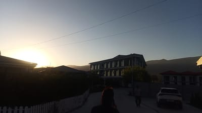 The image shows a street scene in Choktal, Kyrgyzstan, at dusk. A person's back is to the camera, with their head turned to their right, appearing to be on their phone. They are walking on a paved road. To the left, a white picket fence runs along the side of the road, with dark foliage behind it. A house is visible beyond the fence. In the center of the image, a three-story building with numerous windows is situated. To the right, a white SUV is parked, facing away from the camera. Further in the background, mountains are silhouetted against the sky. The sky is clear and transitioning from the bright yellow of sunset to a pale blue. Two power lines are visible stretching across the sky.