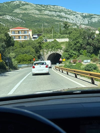 A POV shot from inside a car driving on a road towards a tunnel entrance. A white Volkswagen hatchback with license plate "LO-BI-315" is in front, with a white van following closely behind it. The road has a white dividing line and is bordered by a yellow guardrail on the right. Lush green trees and a mountainous landscape with rocky outcrops dominate the background, with a yellow building with a red roof visible on the left side of the frame. The sky is clear and blue. The scene appears to be set on a sunny day.