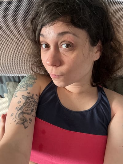 The image shows a close-up, overhead shot of a woman lying down. Her body is angled, with her head to the right and her arm extended to the left. She has dark, wavy hair, brown eyes, and is wearing a two-toned tank top with a navy blue top half and a bright pink bottom half. A tattoo of a creature resembling a mermaid cat is visible on her left arm. The background appears to be a mesh-like fabric, possibly part of a cot or hammock, with some textured pillows visible below it. The setting does not provide enough information to determine a specific location within Červená Píska, Czechia.