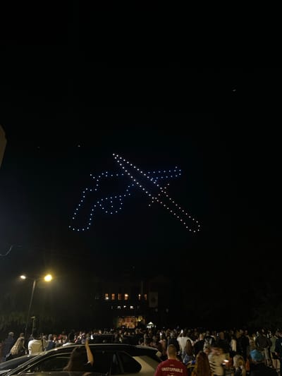 At night in Bratsigovo, Bulgaria, a crowd watches a drone display forming the image of Hristo Prodanov, a Bulgarian mountaineer, against a dark sky. Streetlights illuminate the gathering while building windows are visible in the background. A car and outstretched arms are partially visible on the right.