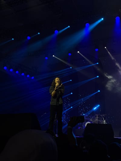 The image depicts an energetic live concert or performance scene, likely at night, in Pagedangan, Indonesia. The setting is dark and atmospheric, dominated by numerous bright blue and some white spotlights that project strong beams of light through visible haze or smoke.
At the center, a person with long dark hair is performing on stage, holding a microphone. Dressed in dark clothing, including a long-sleeved shirt and pants, they appear to be singing passionately, with their mouth open. Blue light from the stage illuminates them from behind and to the side.
The stage infrastructure includes dark metal truss structures overhead and along the sides, supporting the various lights. Large, dark speaker cabinets or other stage equipment are visible on the right, partially obscuring a drum kit, which is also illuminated by blue light. In the foreground on the right, several silhouetted heads suggest audience members or crew are present. One individual's hand is visible holding a smartphone, presumably recording the event. Dark, circular objects, possibly balloons, are also in the extreme foreground. The overall ambiance is vibrant and immersive, characteristic of a live music event. No discernible text is visible within the image.