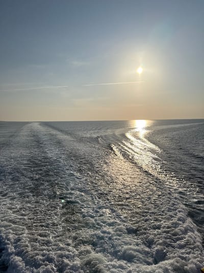 The image depicts a stern view from a vessel underway on the open sea near Palaiopoli, Greece, under a clear sky. The sun, a brilliant point of light, is positioned in the upper right quadrant of the frame, low above the horizon, casting a direct, shimmering path of intense light across the water's surface towards the viewer. The vessel's wake dominates the foreground and midground, presenting a turbulent, foamy trail of churning white water and disturbed dark blue ripples extending towards the distant horizon. Two faint, linear contrails are visible horizontally in the light blue upper sky. No individuals or additional man-made objects are visible within the scene.