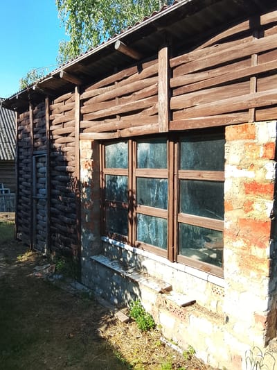 The image shows an exterior view of a rustic wooden structure, possibly a shed or outbuilding, under a clear blue sky with sunlight filtering through tree branches overhead. The building features walls constructed from horizontal wooden logs and planks, with a woven log section on the left side, which appears to be a door or entrance. A large multi-paned window with a wooden frame is visible on the right side, set into a foundation of rough brickwork. The ground around the building is covered in dry grass, dirt, and some sparse greenery. The overall impression is one of a rural or semi-rural setting, consistent with a location in a village or the outskirts of a populated area like Патоцкі сельскі Савет, Belarus. There are no people or animals present in the image.