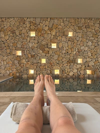 A first-person perspective shot shows a person's legs and feet resting on a lounge chair at a spa in Artà, Spain. The lounge chair has a white cushion and two folded towels, one supporting the person's calves. The person's feet are bare and facing upwards, with toenails painted a light yellow. Their legs are visible, with some bruising on the left leg and a tattoo on the right ankle.

Behind the lounge chair, a pool with dark water is visible, reflecting the light from a stone wall. The wall is constructed from irregular, rough-hewn stones in varying shades of brown and grey. Small, square openings are cut into the wall, each illuminated from within by a warm light, creating a grid-like pattern. The spa appears to be an indoor or partially covered space, with a neutral-colored ceiling visible at the top of the frame.