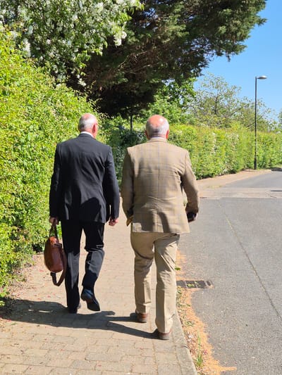 Two men are depicted walking away from the camera on a paved pathway under a clear blue sky in South Cambridgeshire, United Kingdom. The path runs alongside a strip of green foliage, including bushes with white blossoms and tall trees, and an asphalt road.
The man in the foreground, closer to the viewer, is dressed in a dark suit with a white shirt collar visible and is carrying a brown leather briefcase in his left hand. He has a bald head with grey hair on the sides. Slightly behind and to his right is another man, also with a bald head and grey hair, wearing a light brown plaid jacket and light-colored trousers.
The bright sunlight casts distinct shadows of the men on the path. A street light pole with a light fixture is visible against the clear sky in the upper background. Further down the asphalt road, a drain grate is noticeable. The abundant green vegetation and sunny conditions suggest a pleasant outdoor setting. No text is visible in the image.