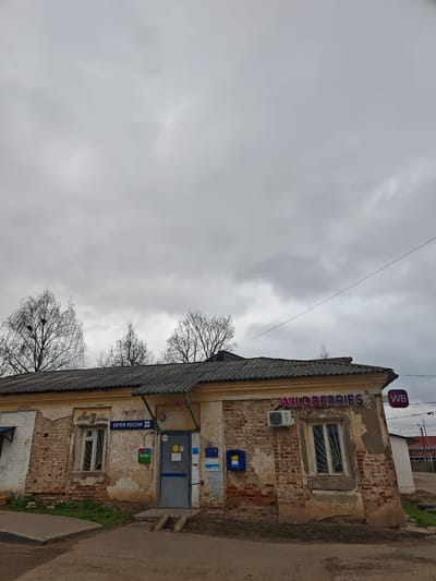 The image captures a dilapidated building in посёлок совхоза "Буденновец", Russia. The structure appears to house both a почта россии (Russian Post Office) and a Wildberries location, indicated by signage. The building is constructed primarily of brick, with extensive crumbling and deterioration of the facade, particularly the plaster. The roof is tiled and exhibits some sagging. Windows are present on both sides of the entrance, covered with bars. A blue mailbox is visible. An Avito sign is next to the entrance. The sky is overcast, and there are bare trees in the background. The area in front of the building appears to be a paved road or parking area. The overall impression is one of neglect and disrepair.