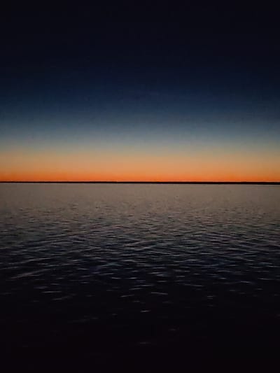 The image captures a view of a body of water at dusk or dawn, with a horizontal horizon line dividing the frame into the sky and the water. The sky transitions from a dark blue at the top to an orange glow near the horizon, suggesting either a sunset or sunrise. The water is dark with subtle ripples, creating patterns of light and shadow. There are no visible people or objects in the water. The location is not specified, so Veregonets, Russia, cannot be confirmed.
