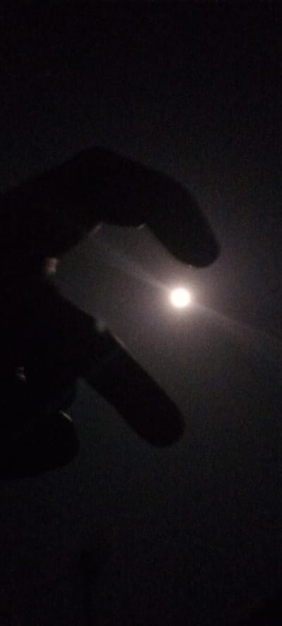 The image depicts a dark, nocturnal scene, likely taken in Ilogbo Eremi, Nigeria. The primary subjects are a human hand, silhouetted in the foreground, and a bright, circular light source, most probably the moon, positioned centrally. The hand is angled with fingers curled, appearing to "hold" or frame the moon, a common photographic gesture. The setting is characterized by profound darkness, indicating nighttime, with the moon serving as the sole significant source of illumination. Its bright glow is visible through the gap formed by the fingers, and a faint halo or lens flare extends from it. The sky appears clear, without visible clouds, allowing for an unobstructed view of the moon. No other objects, landscapes, or background details are discernible due to the low light conditions. There is no visible text in the image. The photograph captures a moment of interaction with the night sky, focusing on the interplay of light and shadow created by the hand and the moon.