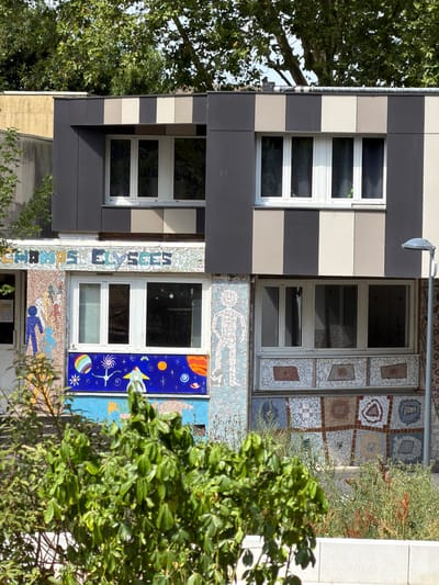 A multi-story building facade is adorned with decorative mosaic tiles and features several windows. The building's exterior is a mix of dark gray and light beige panels arranged in horizontal stripes. On the left side of the image, a window is framed by these striped panels, with a large tree providing a leafy backdrop. Further to the right, another window is surrounded by a detailed mosaic depicting various geometric patterns and abstract shapes. Below this window is a section of mosaic art featuring human figures. To the right of this mosaic, a window is placed next to a vibrant blue mosaic panel decorated with celestial bodies and a rocket ship, suggesting a space or astronomy theme. Another window is visible to the right of this, partially obscured by green foliage. The overall scene suggests an urban setting, possibly a school or cultural center, given the playful and artistic decorations. The time of day appears to be daytime, with bright natural light illuminating the scene. No specific activity or event is evident, but the decorative elements suggest a lively and creative environment. The text "CHAMP ELYSEES" is visible in mosaic lettering along the facade.