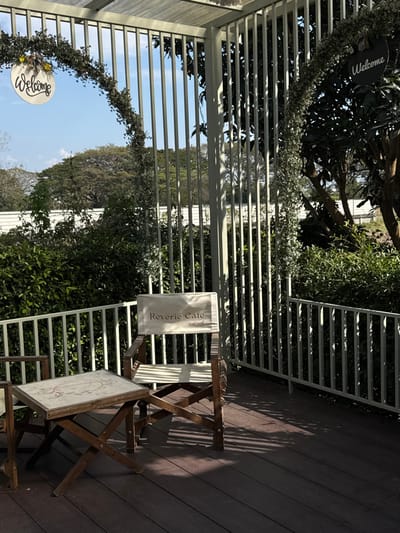 The image displays an unoccupied outdoor seating area of Reverie Cafe, located in San Phra Net, Thailand. The scene is set on a dark wooden deck, across which prominent diagonal shadows are cast by an overhead structure. In the foreground, a light-colored wooden folding table with an indistinct circular design on its top stands between two wooden director's chairs. The chair on the right features a light canvas seat and a backrest labeled "Reverie Cafe" and "เรฟเวอรี่" in black script. A white metal railing with vertical balusters delineates the edge of the deck. Above the railing, a dense line of green bushes and trees is visible, with a white linear structure positioned behind them. The upper portion of the scene is characterized by a white metal pergola-like structure, from which vertical slats descend. Two circular "Welcome" signs, one white with black cursive text on the left and a partially visible black one on the right, are affixed to decorative green floral garlands draped along the structures. The background reveals a clear, light blue sky above a distant treeline. No individuals are present in the scene.