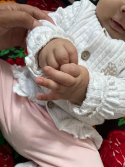A close-up shot shows a baby's hands resting on their lap, wearing a white textured cardigan with buttons. The baby's right hand is curled into a fist, while the left hand is slightly open, with fingers bent. A person's hand is visible near the baby's left arm, gently holding the cardigan. The baby is wearing pink pants and is positioned against a dark fabric with a floral pattern. The scene appears to be indoors, possibly in a home environment in the city of Ак-Сай, Kyrgyzstan.