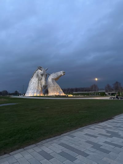 The image displays The Kelpies, two colossal equine head sculptures, illuminated by warm yellow light from their base, against a dark, overcast blue-grey sky. The scene is located in Falkirk, United Kingdom, during evening or twilight hours. The sculptures are composed of numerous metallic panels and stand within a circular water feature or landscaped area. The taller, primary head faces forward and slightly left, while the secondary head is angled downward and to the right. In the foreground, a paved pathway with interlocking light and dark grey rectangular blocks runs alongside a manicured green lawn. In the mid-distance on the right, two individuals are seated at a light-colored wooden picnic table with attached benches. The background includes a line of bare deciduous trees, an elevated road structure, and a bright, distant streetlamp. Faint power lines and industrial structures are visible on the far left horizon.