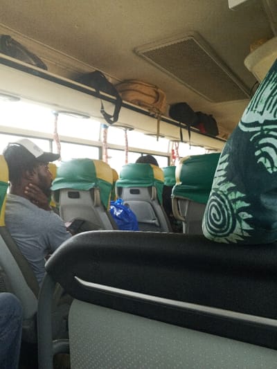 The scene is inside a passenger bus, located in the city of Ibafo, Nigeria. The image captures the interior from a slightly elevated, forward-facing perspective, likely from a seat within the bus.

In the immediate foreground, the top of a black seatback is visible, featuring a white horizontal strip. To its right, a dark green fabric with a light green and white spiral and leaf pattern is partially visible, consistent with a headrest cover.

Further into the bus, a male individual is seated in a grey t-shirt, wearing a white and black baseball cap backward. His right hand is positioned at his neck/chin area. His seat features a grey backrest with a yellow and green fabric cover over the headrest portion. Adjacent to him, a blue plastic bag rests on the floor or a seat. Several rows of similar grey seats with green and yellow headrest covers extend towards the front of the bus. One individual's head is partially visible further back.

Above the seats, an overhead luggage rack runs along the side, holding several bags including a beige backpack and multiple dark-colored bags/backpacks. The bus ceiling is light-colored, featuring a rectangular air vent with a metal grille and several circular light fixtures. Exposed window frames and white-and-red striped fabric loops (possibly handle straps) are visible along the upper window line, indicating ample natural light entering the vehicle.