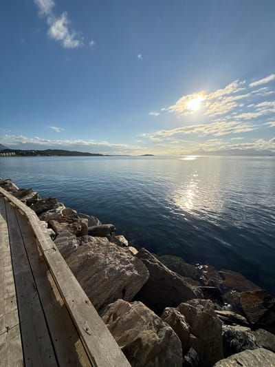 The image depicts a scenic view of the sea with a wooden boardwalk and a rocky shoreline in the foreground. The sun is shining brightly in the sky, casting a reflection on the water. In the distance, a coastline with hills and buildings can be seen. The location is identified as Harstad, Norway. There are no people or animals visible in the image. The scene is peaceful and serene, with the natural beauty of the landscape being the main focus. The boardwalk appears to be made of weathered wood, and the rocks are rough and uneven, suggesting a natural and unmanicured environment. The water is calm and clear, with gentle ripples reflecting the sunlight. The sky is a clear blue with scattered white clouds. The overall impression is one of a tranquil and picturesque coastal setting.