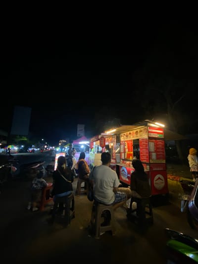 This is a nighttime, eye-level, medium shot of a busy street food scene in Tanggul, Indonesia. The primary focus is on a food stall with a red and white exterior, illuminated by bright lights. The stall displays various menu items, including what appears to be "Kotaki" and "GO FOOD" branding. Several people are gathered around the stall. On the right, a person wearing a yellow hoodie and a pink skirt is visible. In the foreground, a man in a white t-shirt and checkered shorts is seated on a stool, facing away from the camera. To his left, a woman wearing a dark hijab and brown jacket is also seated. Further left, a child in a patterned outfit is standing at a small red table. In the background, other food stalls with colorful umbrellas and tents line the street, and motorbikes are parked along the side of the road, indicating a lively night market atmosphere. The dark sky suggests it is late at night, with some artificial lighting from the stalls and distant streetlights creating an ambient glow.