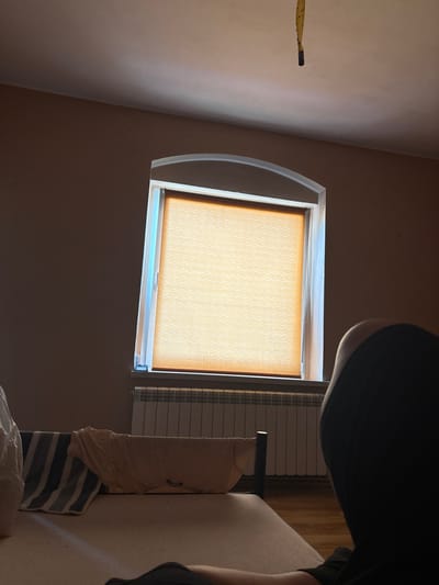 The image depicts an indoor scene, likely a bedroom, with a arched window covered by a light orange roller blind. Sunlight streams through the sides of the blind, creating a soft illumination. Below the window, a white radiator with multiple horizontal panels is visible. In the foreground, a bed is partially seen, with a striped towel and a beige cloth draped over its edge. To the right, a person's dark silhouette is prominent, with their back and head facing away from the camera. The room's walls are painted a neutral, light brown color. A yellow flypaper strip with caught insects hangs from the ceiling in the upper part of the image. The flooring appears to be wood. This scene is located in Słupia pod Kępnem, Poland.