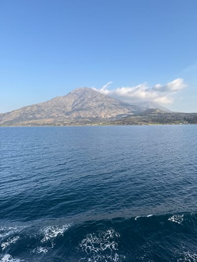 The image presents a wide, expansive view of the coastal landscape of Kamariotissa, Greece. A prominent, rugged mountain range, identified as Mount Saos, dominates the background, displaying grey, rocky terrain with scattered patches of dry vegetation. The peaks on the right side of the range are partially obscured by scattered white cumulus clouds, contrasting with the otherwise clear, bright blue sky above. Along the base of the mountain, directly at the shoreline, small clusters of white and light-colored buildings are visible, identifying the city of Kamariotissa. The foreground is occupied by a large expanse of deep blue sea, its surface showing distinct ripples and texture. A white, foamy wake is present in the immediate lower left portion of the frame, indicating the photograph was captured from a moving vessel on the water. No individuals or specific actions are discernible beyond the implied movement of the viewpoint.