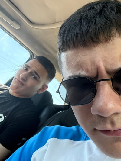 The image appears to be a selfie taken inside a car. Two men are visible. One is closer to the camera, wearing sunglasses, and his face is partially in the frame. The other man is further back, wearing a black t-shirt and looking at the camera. The interior of the car is visible, including the ceiling, window, and seat. The specific location in the city of Belovitsa, Bulgaria cannot be determined from the image. There is no information given in the image as to location, time or specifics of events.