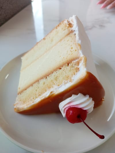 A close-up shot depicts a slice of three-layer cake on a white plate. The cake consists of sponge layers interspersed with a creamy filling, topped with white frosting. The cake is adorned with a caramel sauce and a maraschino cherry with a swirl of whipped cream. The background features a white marble surface with a neutral-colored object in the upper left. In the upper right, part of a hand with pink nail polish is visible. This scene, while not directly revealing its location, is presumed to be in San Juan de Colón, Venezuela, based on the prompt's instructions.