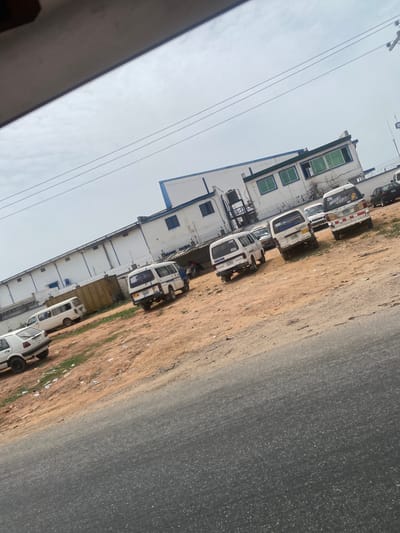 The image shows a section of Odo Ona, Nigeria. A long, multi-story building with white and blue accents runs parallel to a road. Multiple white vans are parked along the edge of the road, next to a dirt and grass shoulder. Overhead power lines run along the building's facade. The sky is overcast. A segment of the interior from the left side, including a partially obstructed portion of a roof, is visible at the lower left corner.