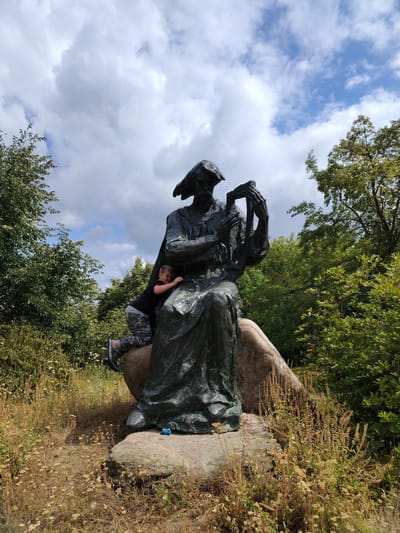 In Balyko-Shchuchynka, Ukraine, a young boy is pictured climbing on a large bronze statue situated outdoors amongst lush greenery and a boulder. The statue depicts a figure seated with a harp-like instrument. The boy, wearing camouflage pants and a black shirt, is positioned on the side of the statue, his body contorted as he grips the sculpture for support. The surrounding environment is wild, with dry grasses and wildflowers growing around the base of the statue and boulder. Trees and foliage frame the scene, with a cloudy sky overhead. A small blue toy is visible on the boulder at the base of the statue.
