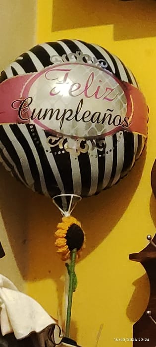 A celebratory birthday balloon with black and white stripes is the focal point. "Feliz Cumpleaños" is printed on its front in a swirling silver script, framed by a pink ribbon. The balloon is tied with string, which is obscured by a small sunflower. The background is a bright, solid yellow wall. A partial view of an ornate wooden decoration hangs on the wall. A timestamp is faintly visible in the top right corner.