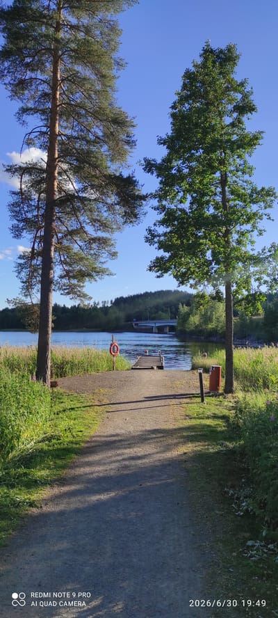 This media file shows a picturesque scene of a canal or riverbank in Joutsa, Finland, on a sunny day. Two large trees, one deciduous and one coniferous, frame the view, with lush green grass and foliage growing along the bank. A pathway, possibly gravel or dirt, runs along the right side of the canal, bordered by more grass.
In the middle ground, a wooden dock extends into the water, and a red life preserver is visible on a post near the bank, suggesting a recreational area. A trash can is also present near the pathway. The water itself appears calm and reflects the blue sky and surrounding greenery. In the distance, a structure that looks like a lock or a bridge crosses the water, with a line of trees forming a dark green backdrop on the far bank.
The time of day appears to be late afternoon or early evening, indicated by the long shadows cast by the trees and the warm sunlight. The weather seems clear and pleasant. The overall atmosphere is peaceful and serene, evoking a sense of tranquility in nature.
Visible text in the media file includes a timestamp and device information on the lower right side, reading "2026/6/30 19:49" and "REDMI NOTE 9 PRO AI QUAD CAMERA".